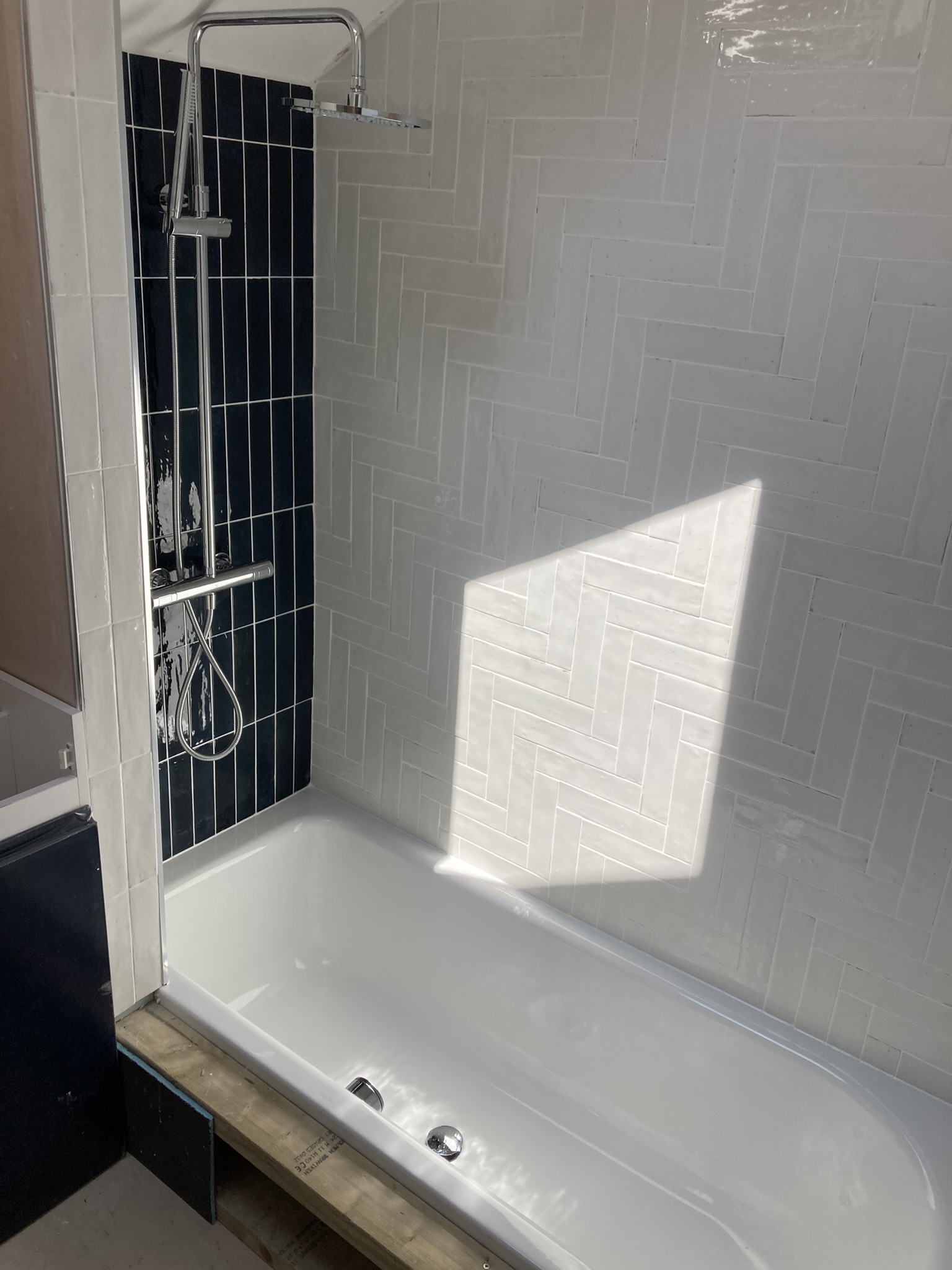 Mid-fit: white herringbone tiling and a black vertical-tile shower recess over a new bath — the tiling taken to the same standard as the plumbing.