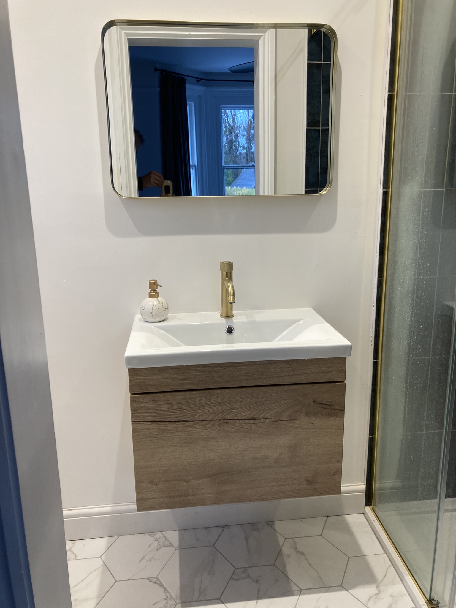 An en-suite with a floating oak vanity, brushed-brass tap, framed mirror and marble hexagon floor beside a glazed shower.